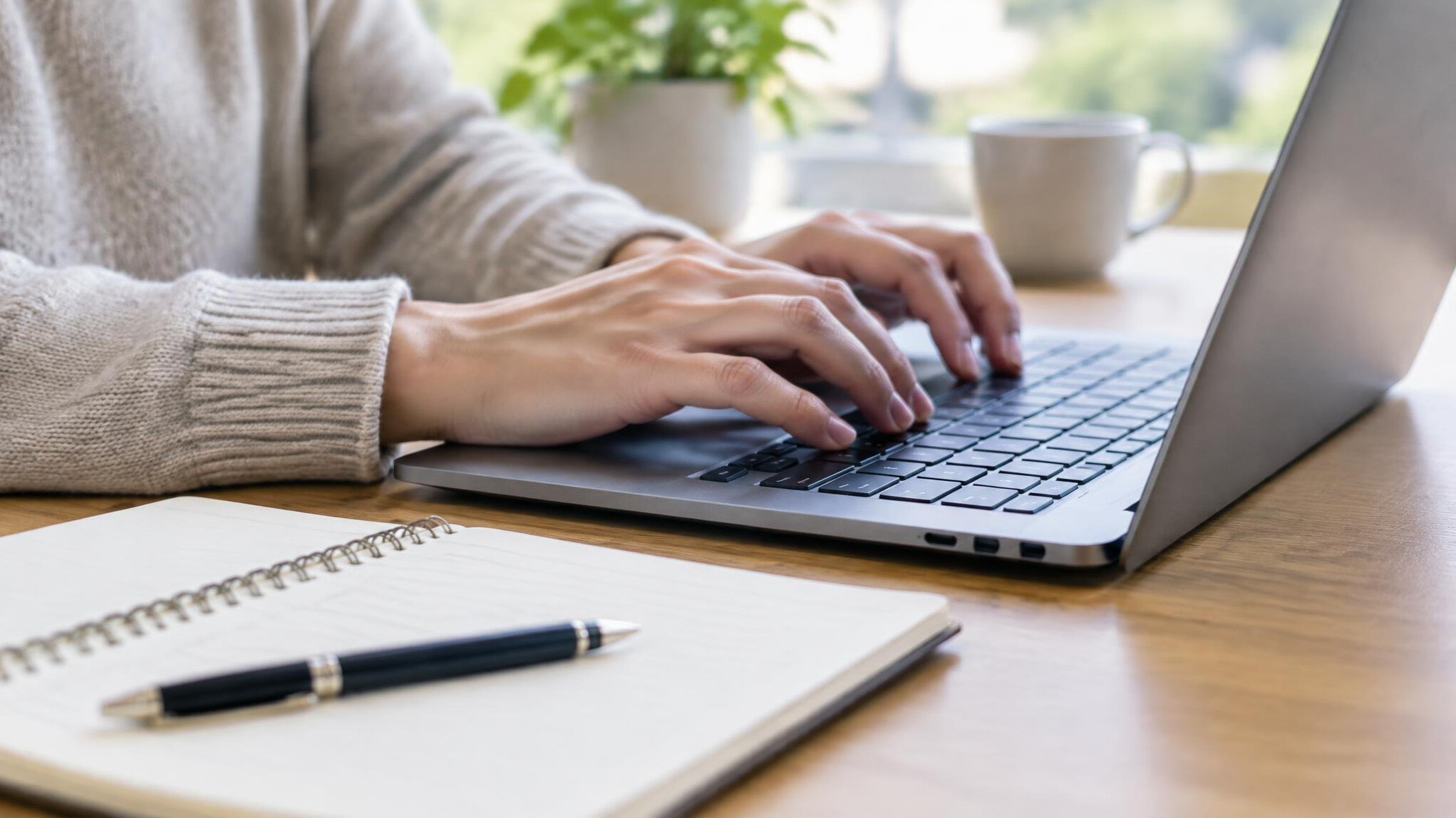 Hands typing on a laptop next to a notebook on a desk